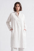 A woman wearing a long white linen tunic with a mandarin collar and front pockets, paired with matching white trousers, photographed against a seamless white studio background.