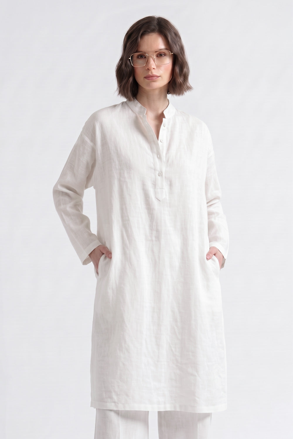 A woman wearing a long white linen tunic with a mandarin collar and front pockets, paired with matching white trousers, photographed against a seamless white studio background.