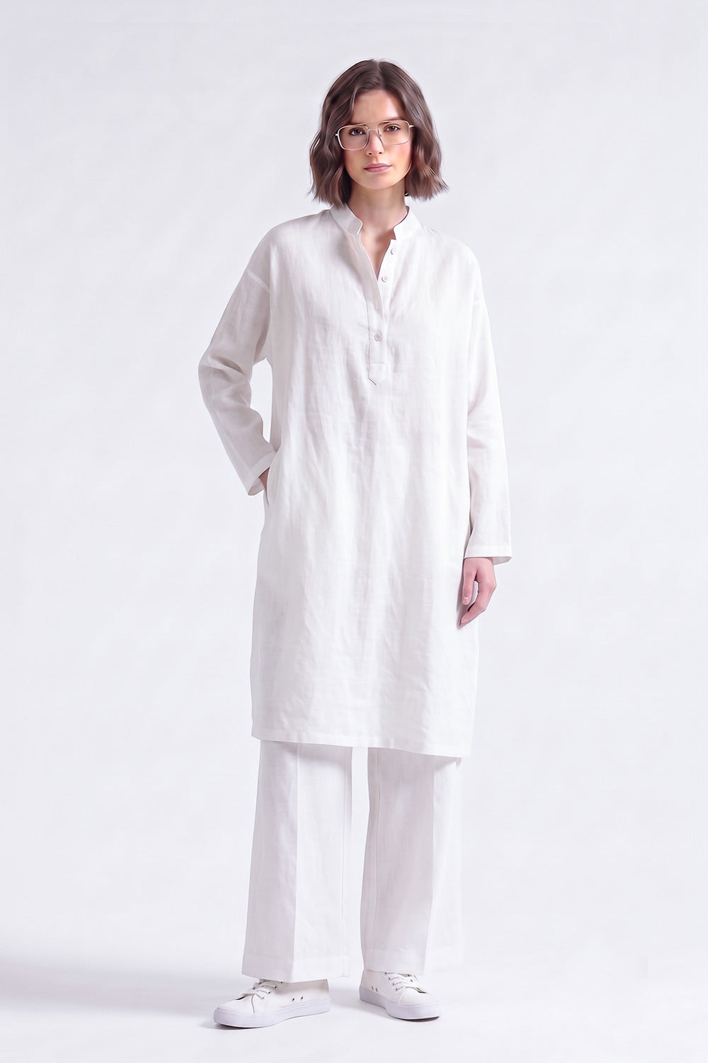 A woman modeling a white long-sleeve linen tunic with a buttoned mandarin collar, paired with matching white trousers and white sneakers, against a plain white studio background.