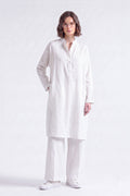 A woman modeling a white long-sleeve linen tunic with a buttoned mandarin collar, paired with matching white trousers and white sneakers, against a plain white studio background.