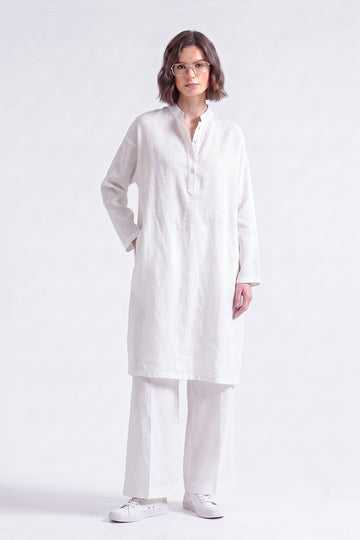 A woman modeling a white long-sleeve linen tunic with a buttoned mandarin collar, paired with matching white trousers and white sneakers, against a plain white studio background.