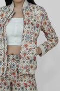 A woman models a floral linen bomber jacket with a collar, featuring a front zipper and ribbed cuffs. She is wearing a white top and white shoes, standing against a plain background.