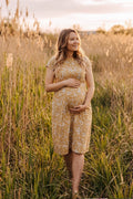 A pregnant woman stands in a field of tall golden grass at sunset, wearing a short-sleeve mustard yellow maternity dress with a white floral pattern.