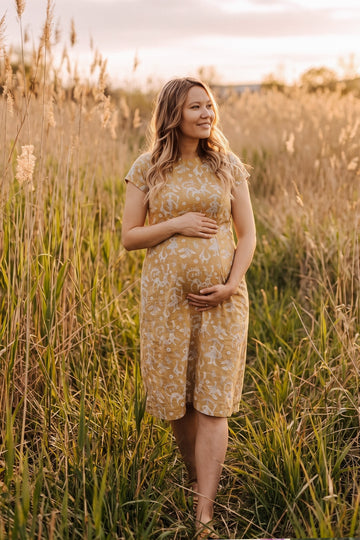 A pregnant woman stands in a field of tall golden grass at sunset, wearing a short-sleeve mustard yellow maternity dress with a white floral pattern.