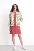 A woman stands against a plain white studio wall wearing a cream bomber-style jacket with a green leaf eco-print over a dusty rose dress, white sneakers, and holding sunglasses.