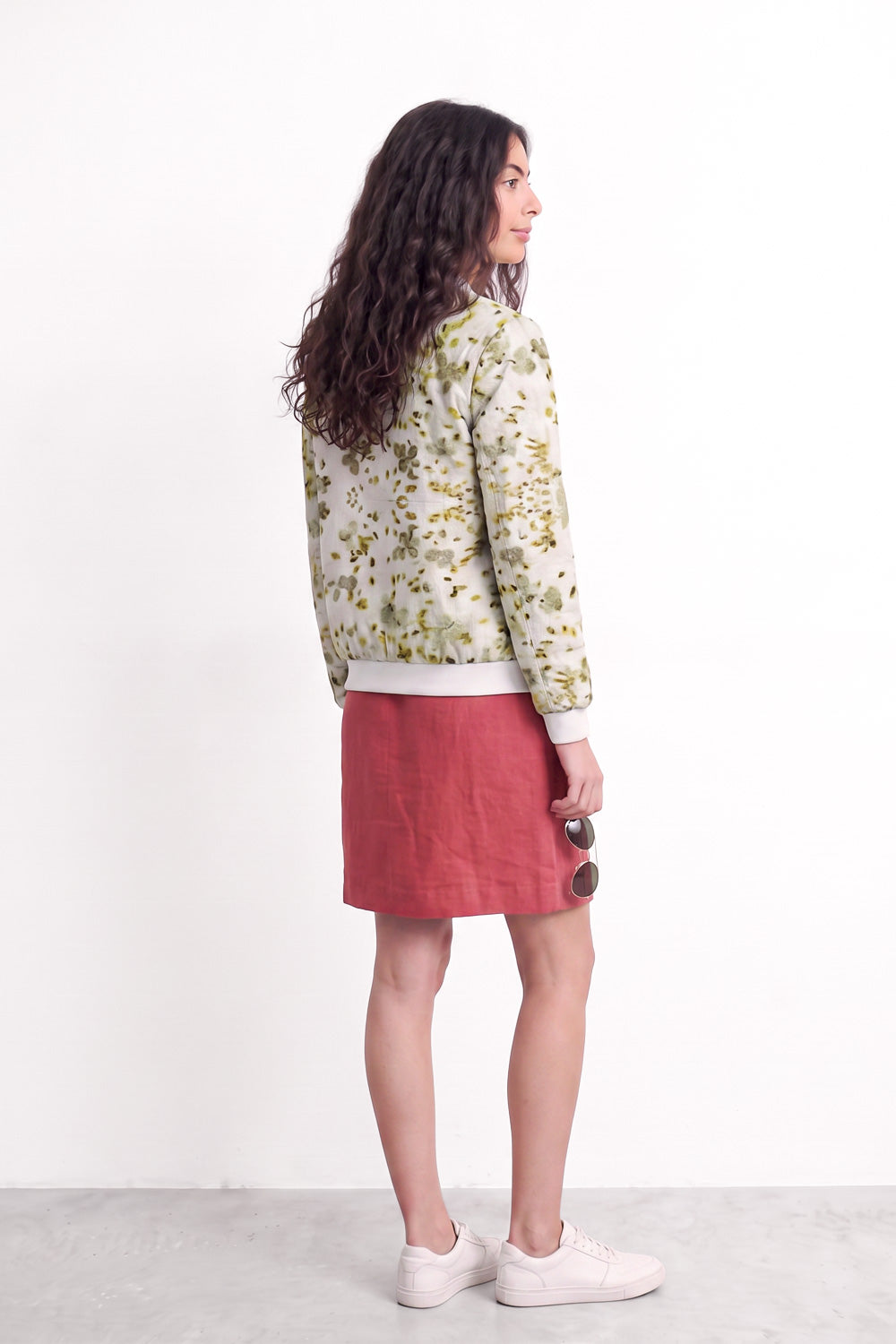 A woman stands in profile wearing a cream-colored long-sleeve bomber jacket with a green and yellow floral eco-print, paired with a dusty rose pink linen mini skirt and white sneakers, against a plain white studio background.
