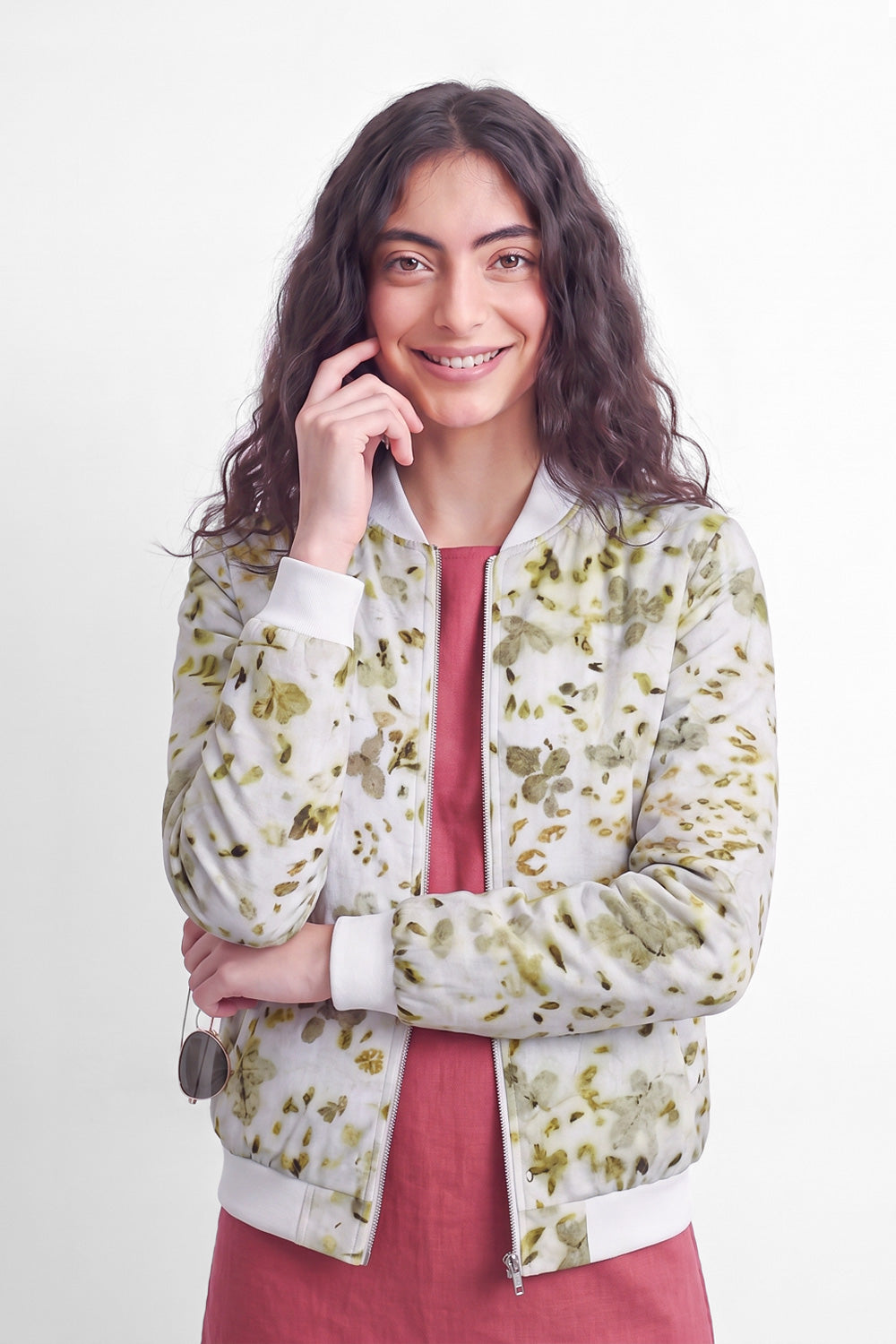 A woman with long dark curly hair models a white bomber jacket with a green and yellow leaf eco-print pattern, worn over a dusty rose dress against a plain white background.