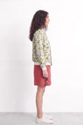 A woman stands in profile wearing a cream-colored bomber jacket with a green and yellow floral eco-print, a dusty rose pink mini skirt, and white sneakers against a plain white studio background.