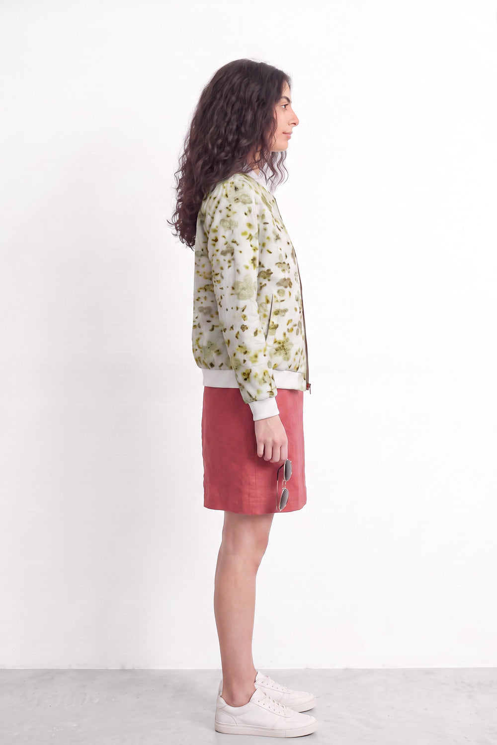 A woman stands in profile wearing a cream-colored bomber jacket with a green and yellow floral eco-print, a dusty rose pink mini skirt, and white sneakers against a plain white studio background.