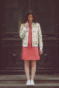 A woman stands on wooden steps in front of a dark carved wooden door, wearing a white bomber jacket with a green floral eco-print, a coral pink dress, and white sneakers.