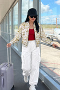 A woman walking through an airport terminal wearing a cream floral eco-printed bomber jacket, white cargo pants, a red top, and a black cap, while holding a lavender suitcase.