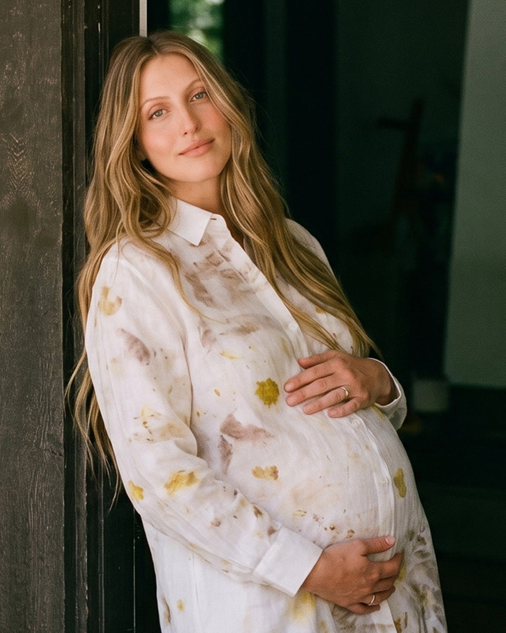 A pregnant woman with long blonde hair leans against a dark wooden door, wearing a white long-sleeve button-up shirt with a subtle gold and brown abstract floral eco-print pattern.