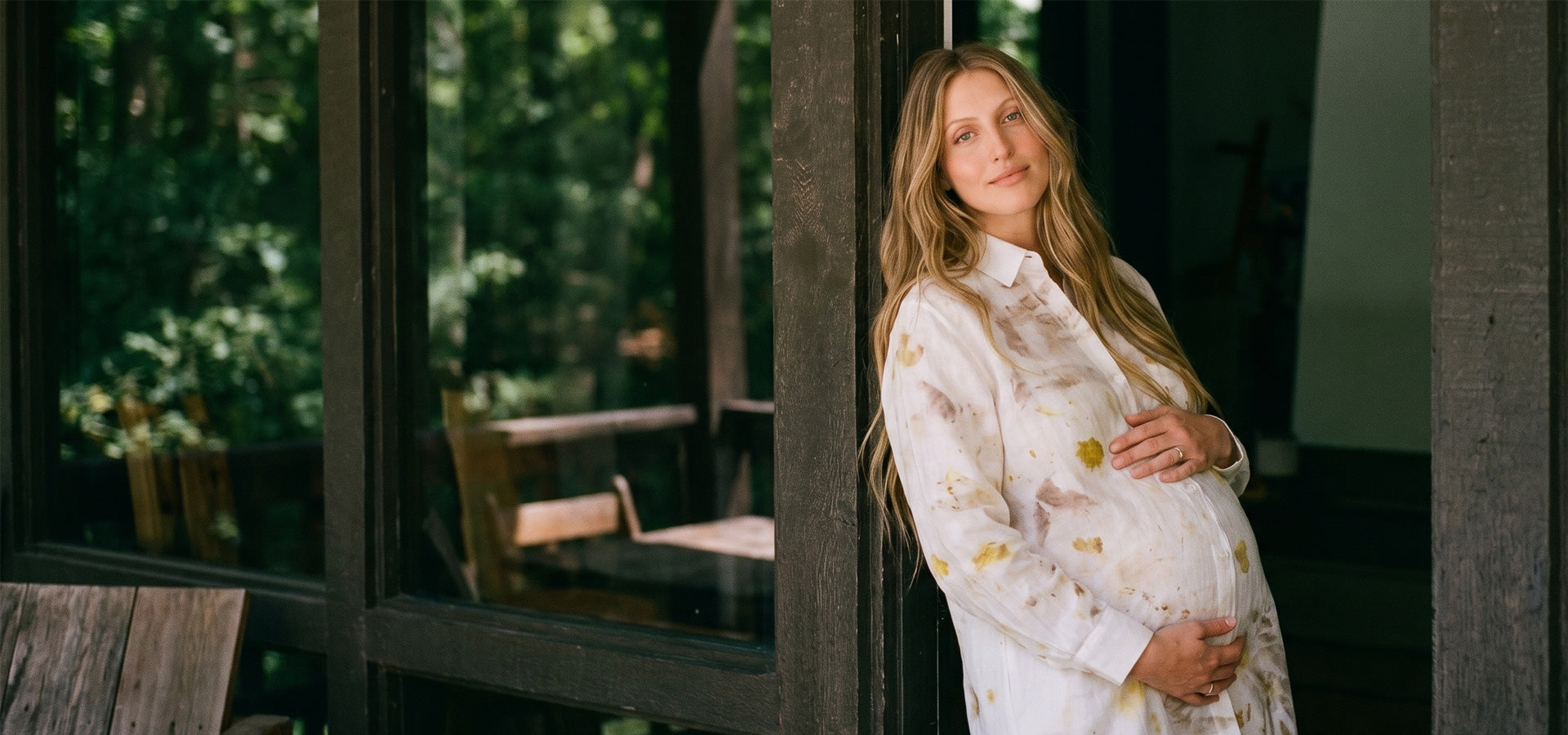 A pregnant woman with long blonde hair stands in a doorway wearing a white long-sleeve button-up shirt with a delicate yellow floral eco-print pattern. She is holding her baby bump, with a rustic wooden deck and lush green forest visible in the background.