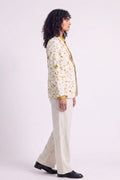 A model shown in profile wearing a cream-colored hemp blazer with a yellow and brown floral eco-print, paired with cream trousers and black shoes, against a light studio background.