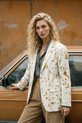 A woman with curly blonde hair stands beside a vintage brown car, wearing a cream-colored hemp blazer with a yellow and green eco-print, a dark green shirt, and beige corduroy trousers, set against a rusty orange wall.