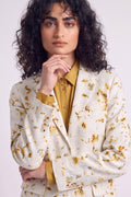 A model with curly dark hair wearing a cream-colored hemp blazer with a yellow and green floral eco-print, layered over a mustard yellow button-down shirt, against a plain light gray background.