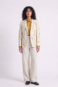 A model stands against a light gray studio background wearing a cream-colored hemp blazer with a yellow and brown eco-print, a mustard yellow collared shirt, cream wide-leg trousers, and black shoes.