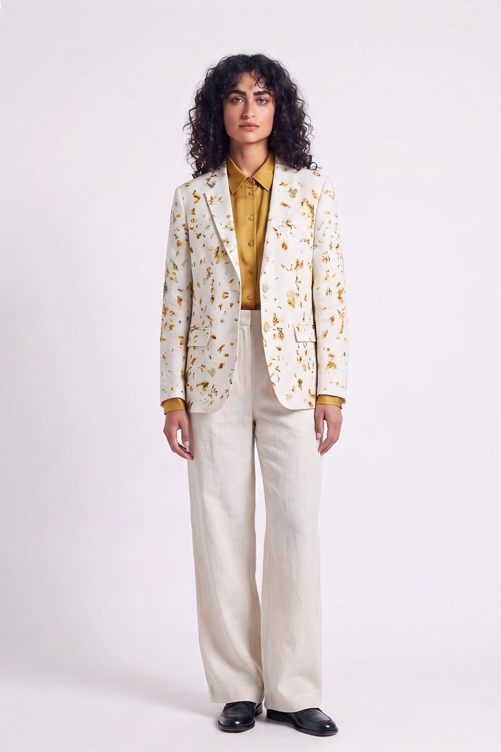 A model stands against a light gray studio background wearing a cream-colored hemp blazer with a yellow and brown eco-print, a mustard yellow collared shirt, cream wide-leg trousers, and black shoes.