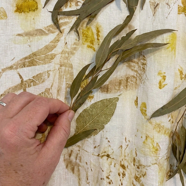 A hand holding a dried bay leaf sprig over a white fabric background with gold leaf pressed botanical designs.