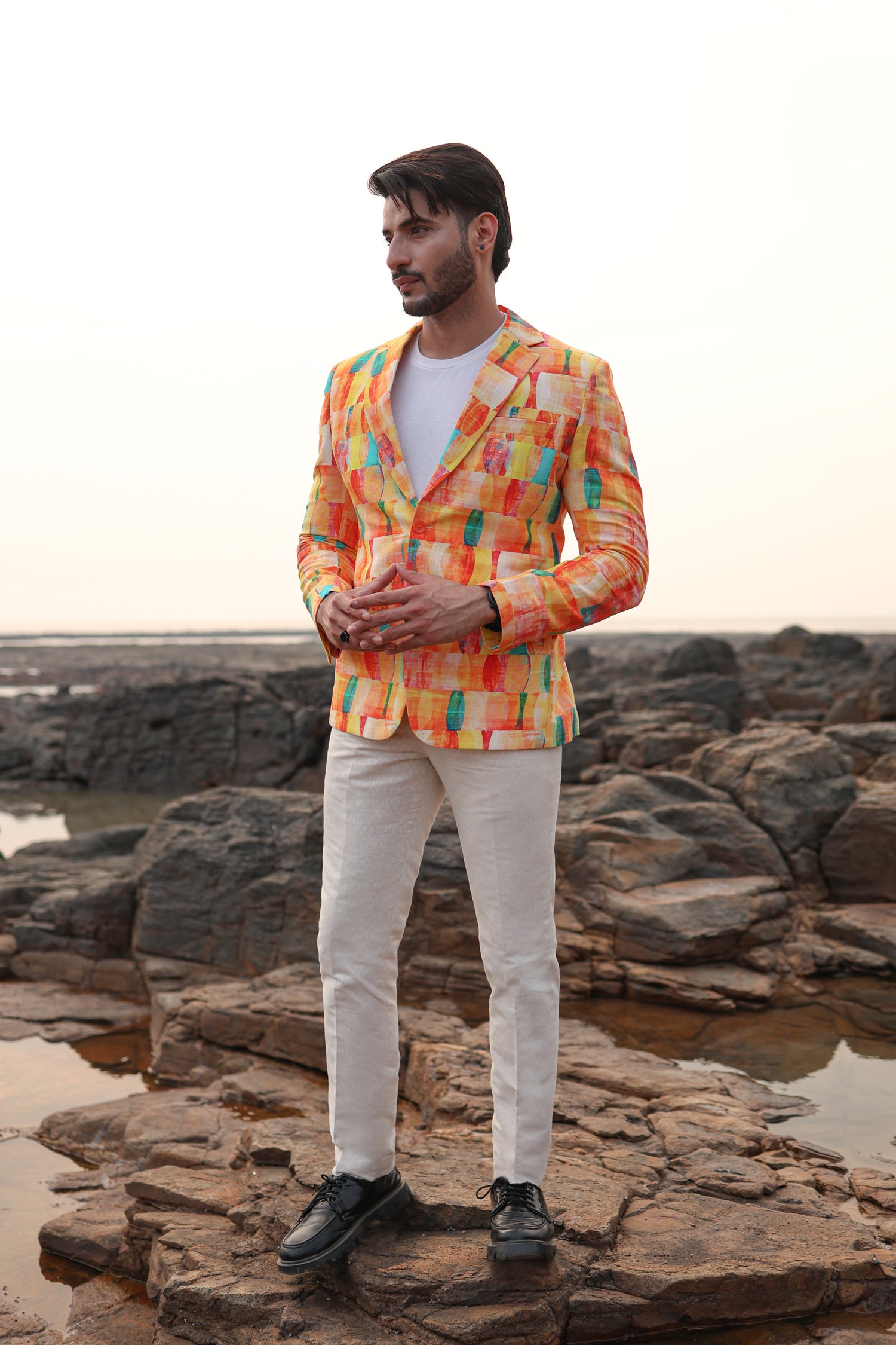 Man wearing a colorful jacket and white pants standing on rocks by a body of water.