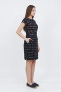 A woman modeling a short-sleeve knee-length linen  dress with a black base and a colorful geometric pattern of small red, blue, yellow, and green squares, paired with dark slip-on shoes against a white studio background.
