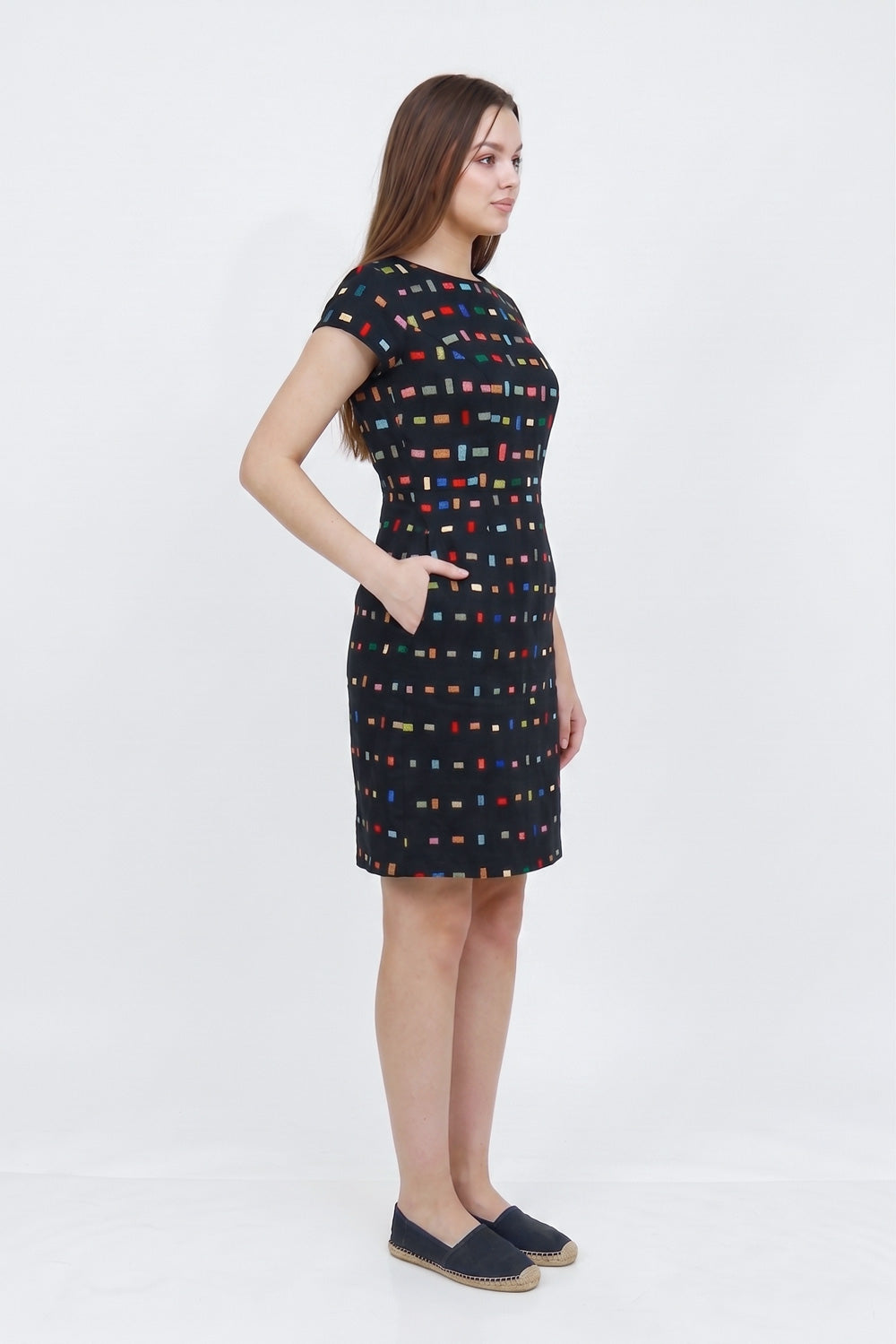 A woman modeling a short-sleeve knee-length linen  dress with a black base and a colorful geometric pattern of small red, blue, yellow, and green squares, paired with dark slip-on shoes against a white studio background.