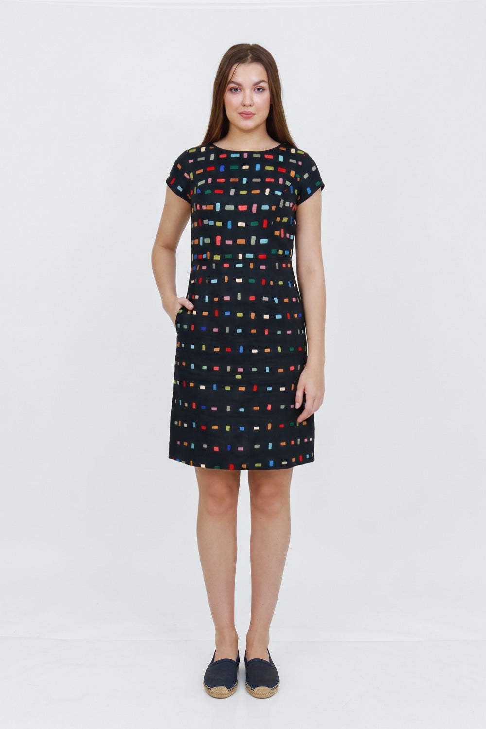 A woman modeling a short-sleeve knee-length dress with a black base and a colorful geometric grid pattern, paired with dark slip-on shoes, against a plain white studio background.