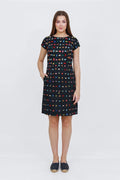 A woman modeling a short-sleeve knee-length dress with a black base and a colorful geometric grid pattern, paired with dark slip-on shoes, against a plain white studio background.