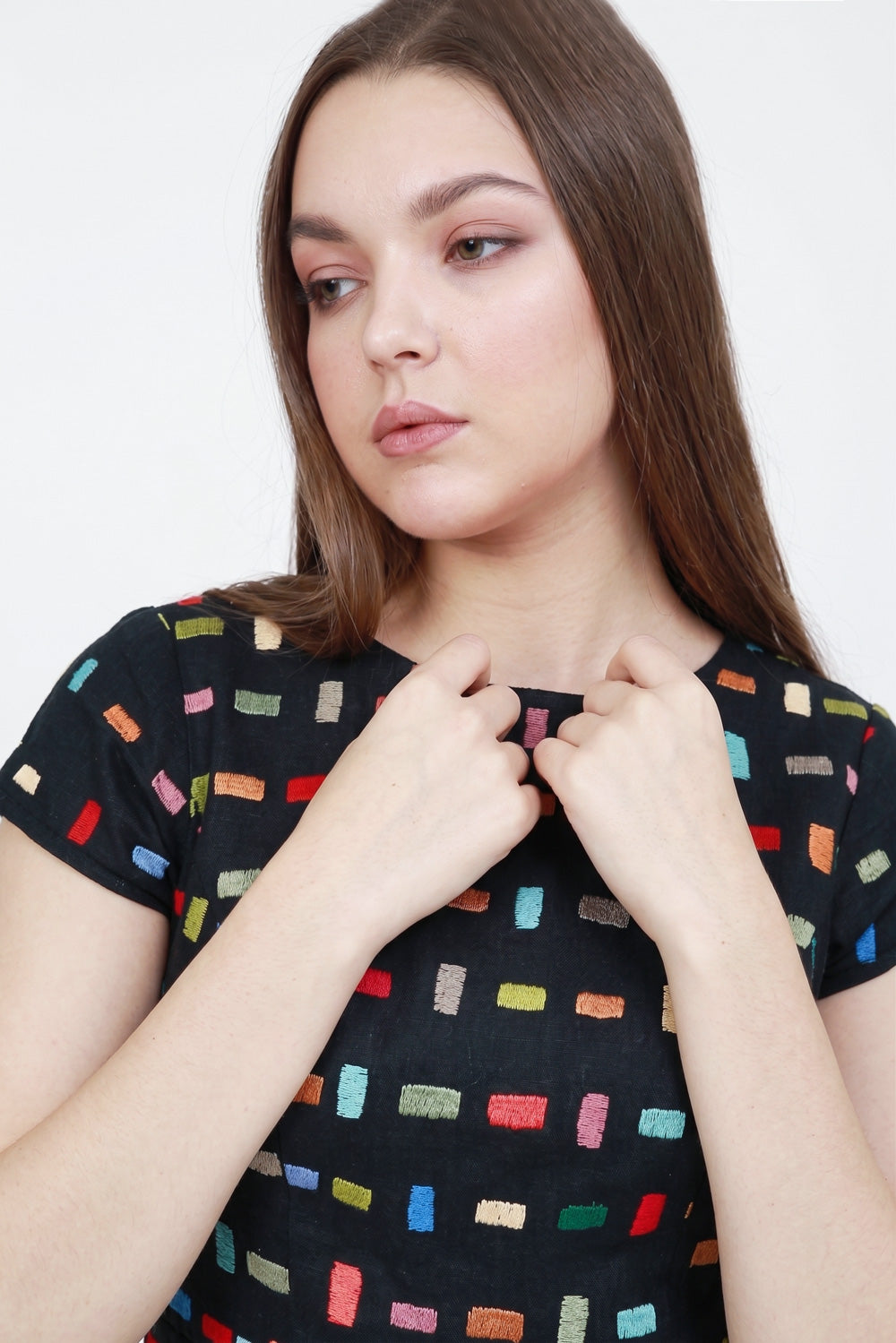 A woman wearing a black short-sleeve tlinen dress with a colorful embroidered geometric pattern of small rectangles, posing with her hands near the collar.
