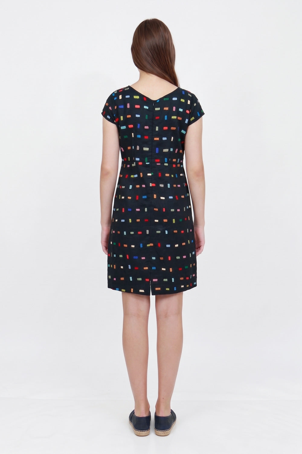 A woman viewed from the back wearing a short-sleeve V-neck linen dress with a multicolor geometric pattern on a black background, paired with dark espadrille flats, against a plain white studio background.