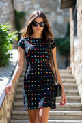 A woman wearing a black short-sleeve shift linen dress with a colorful geometric dot pattern walks down outdoor stone steps. She is wearing black sunglasses and carrying a small black shoulder bag.
