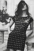 A woman wearing a black short-sleeve mini linen dress with a white geometric grid pattern, posed outdoors against a stone balustrade.