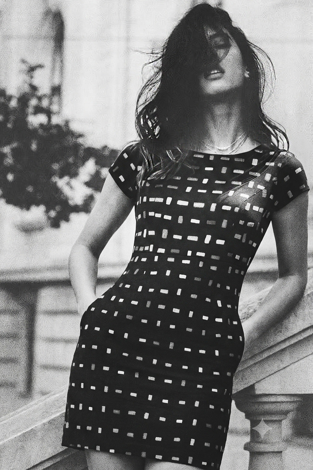 A woman wearing a black short-sleeve mini linen dress with a white geometric grid pattern, posed outdoors against a stone balustrade.
