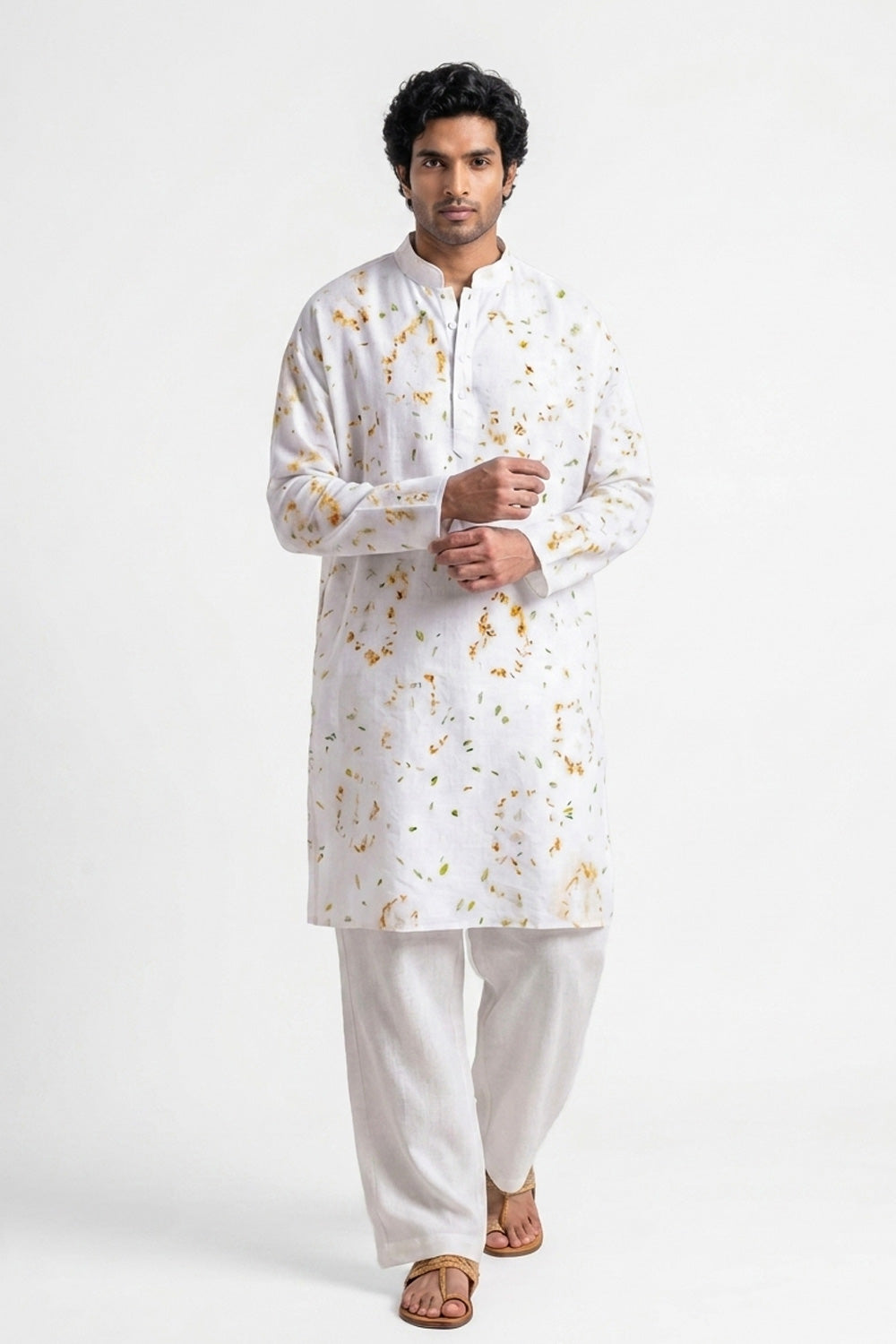 A man modeling a white long-sleeve kurta with a subtle gold floral pattern, paired with white trousers and brown sandals, against a plain white studio background.