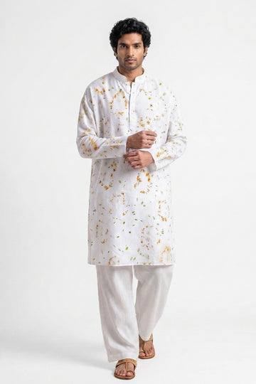 A man modeling a white long-sleeve kurta with a subtle gold floral pattern, paired with white trousers and brown sandals, against a plain white studio background.