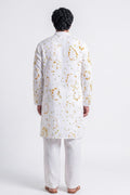 A man viewed from the back wearing a white kurta with a gold abstract pattern and white trousers, standing against a light gray studio background.