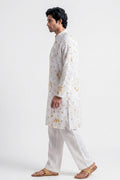 A man in profile wearing a white long kurta with a delicate yellow and green floral print, paired with white trousers and brown leather sandals, set against a plain white studio background.
