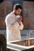 A man in a white long-sleeve kurta with yellow floral patterns stands in a prayer pose in front of a wooden incense burner with burning incense sticks.