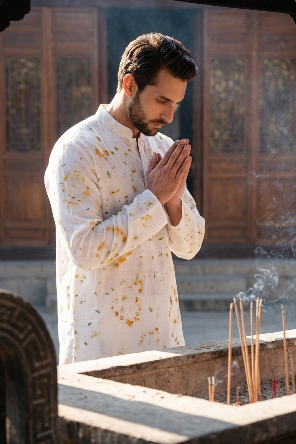 A man in a white long-sleeve kurta with yellow floral patterns stands in a prayer pose in front of a wooden incense burner with burning incense sticks.