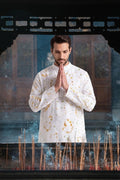 A man in a white long-sleeve shirt with gold tie-dye patterns stands with his hands pressed together in a prayer pose, framed by a dark wooden structure with incense sticks in the foreground.