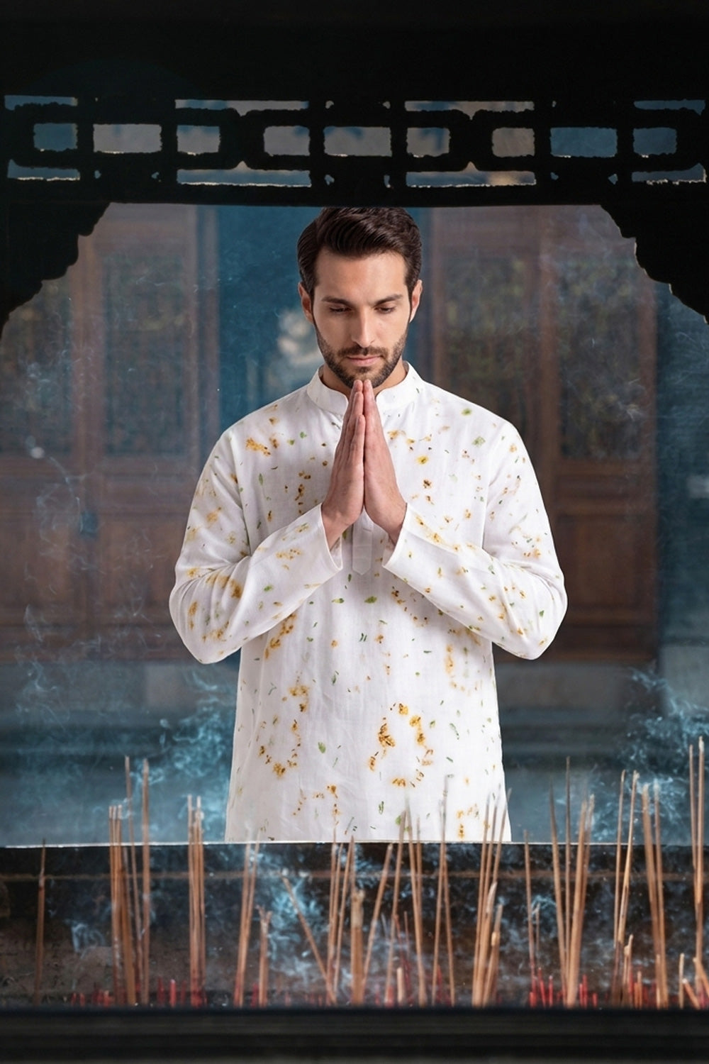 A man in a white long-sleeve shirt with gold tie-dye patterns stands with his hands pressed together in a prayer pose, framed by a dark wooden structure with incense sticks in the foreground.