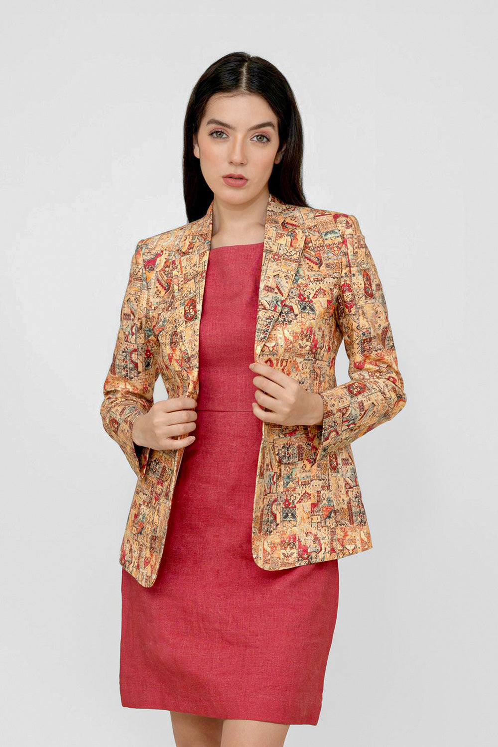 A woman modeling a tailored linen blazer with an intricate multicolor baroque-style print over a solid deep red dress, set against a plain light gray studio background.