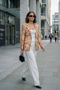 A woman wears a patterned linen blazer in warm earthy tones over a white camisole and wide-leg trousers, styled with black pointed shoes, sunglasses, and a structured handbag while walking through a modern city street.