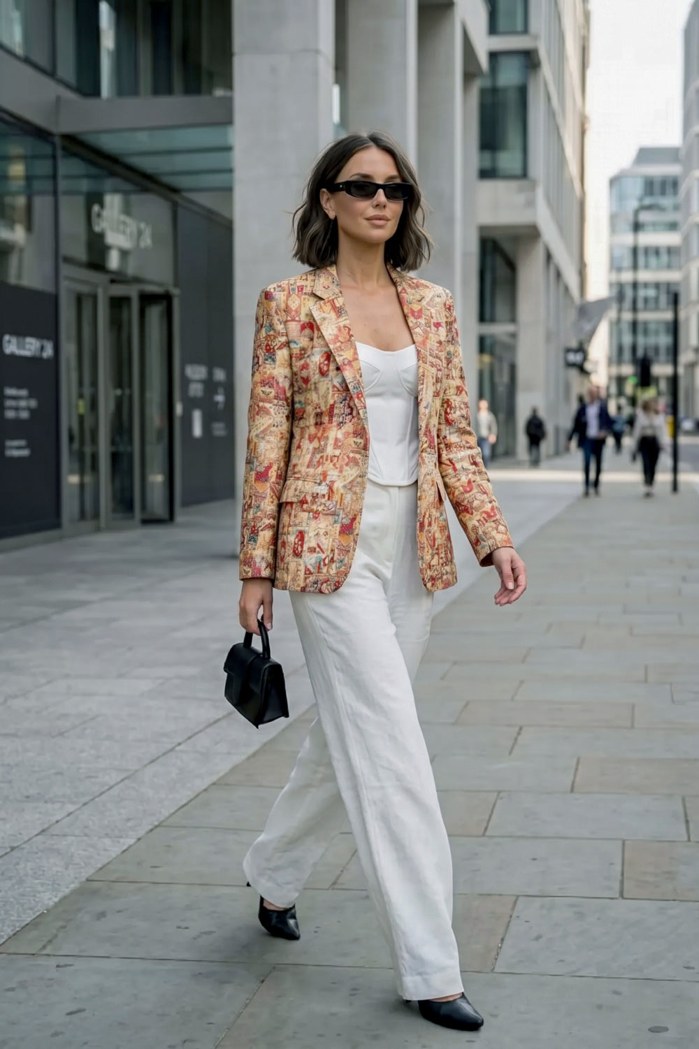 A woman wears a patterned linen blazer in warm earthy tones over a white camisole and wide-leg trousers, styled with black pointed shoes, sunglasses, and a structured handbag while walking through a modern city street.