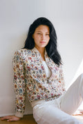 A woman with long black hair sits against a white wall wearing a cream-colored bomber linen jacket with a dense multicolor floral pattern, paired with white pants.