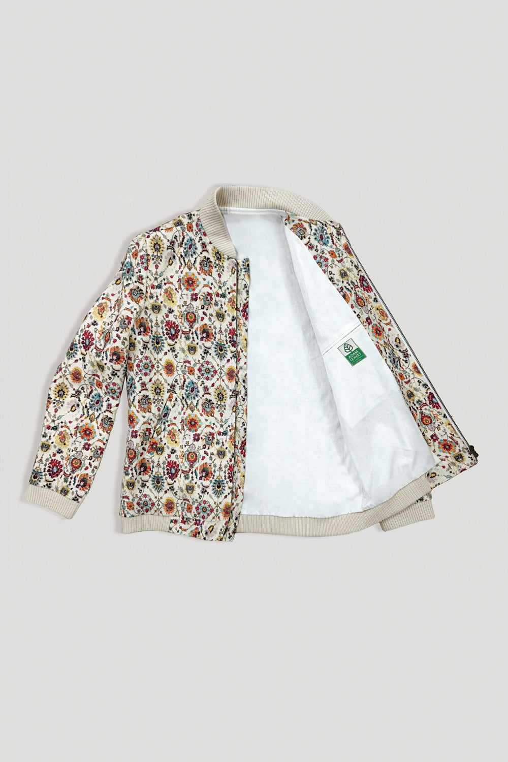 A model stands against a plain light gray wall wearing a cream-colored bomber linen jacket with a dense multicolor floral and botanical print, a white t-shirt, loose-fit blue jeans, and white sneakers.