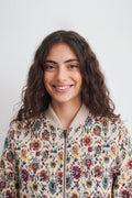 A smiling woman with long, wavy dark brown hair wearing a cream-colored bomber linen jacket with a vibrant, multicolored floral pattern, set against a plain light gray background.