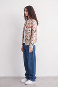 A woman stands in profile wearing a cream-colored bomber linen jacket covered in a dense multicolor floral and botanical print, paired with wide-leg blue jeans and white sneakers, against a plain white studio background.