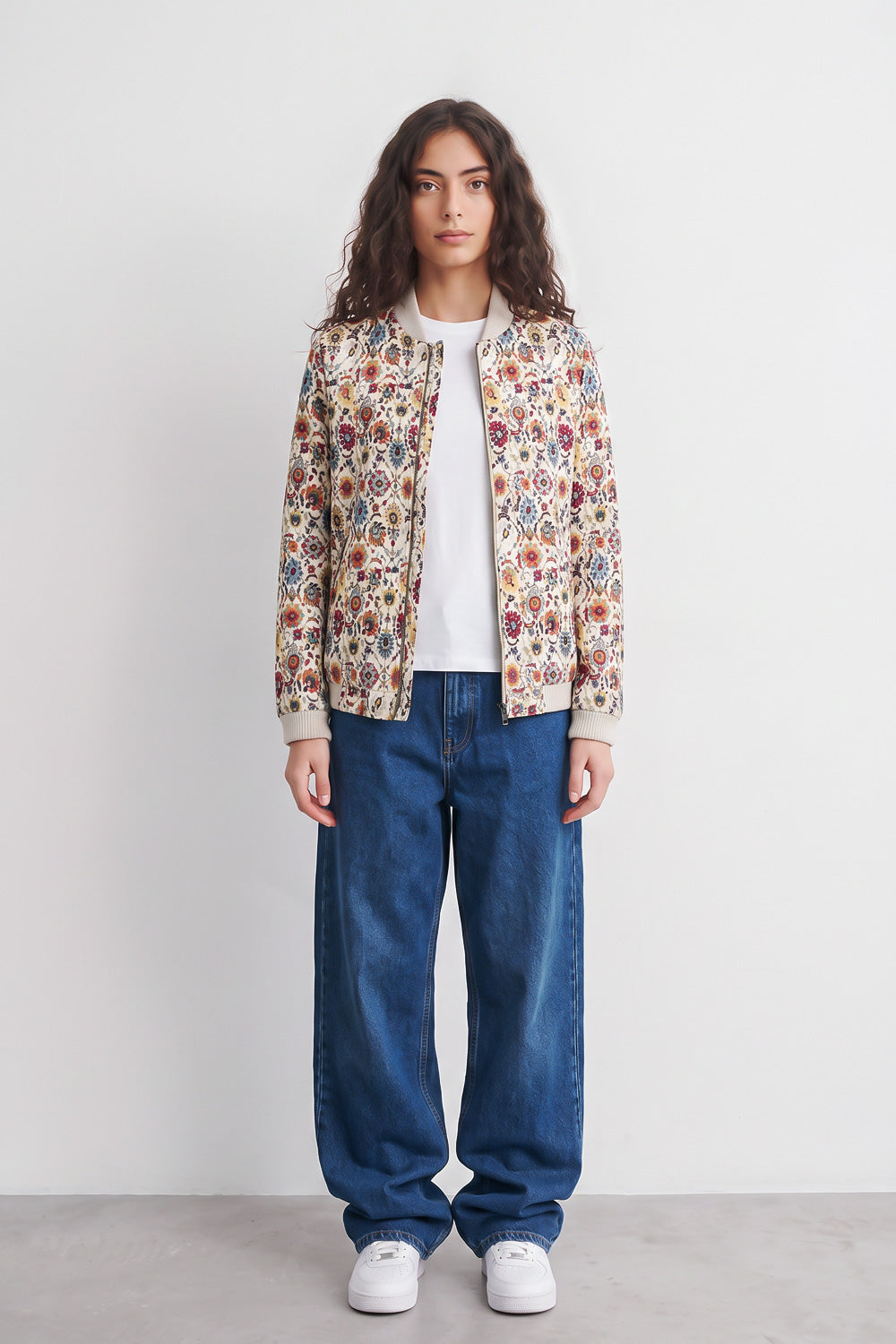 A model stands against a plain light gray wall wearing a cream-colored bomber linen jacket with a dense multicolor floral and botanical print, a white t-shirt, loose-fit blue jeans, and white sneakers.