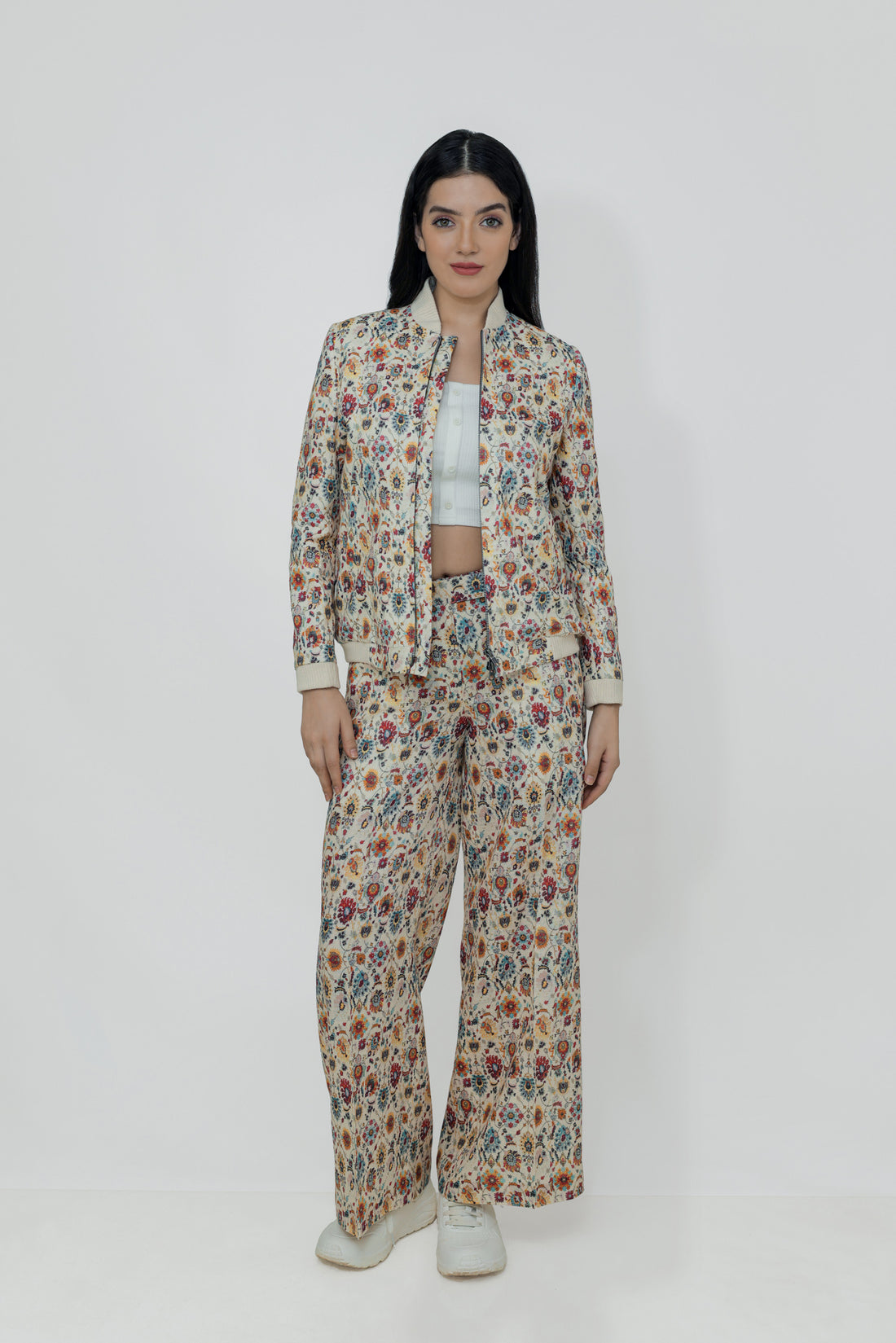 A woman models a floral linen bomber jacket with a collar, featuring a front zipper and ribbed cuffs. She is wearing a white top and white shoes, standing against a plain background.